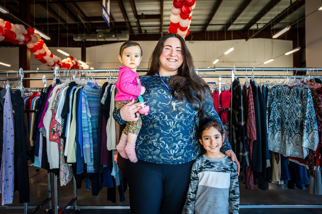 A mother is with her two children, holding one child in front of a clothing rack in the Resource Center.