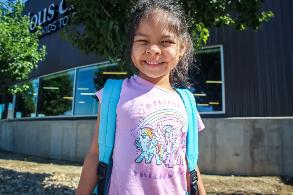 A little girl wearing a unicorn shirt, smiling with her new blue backpack.
